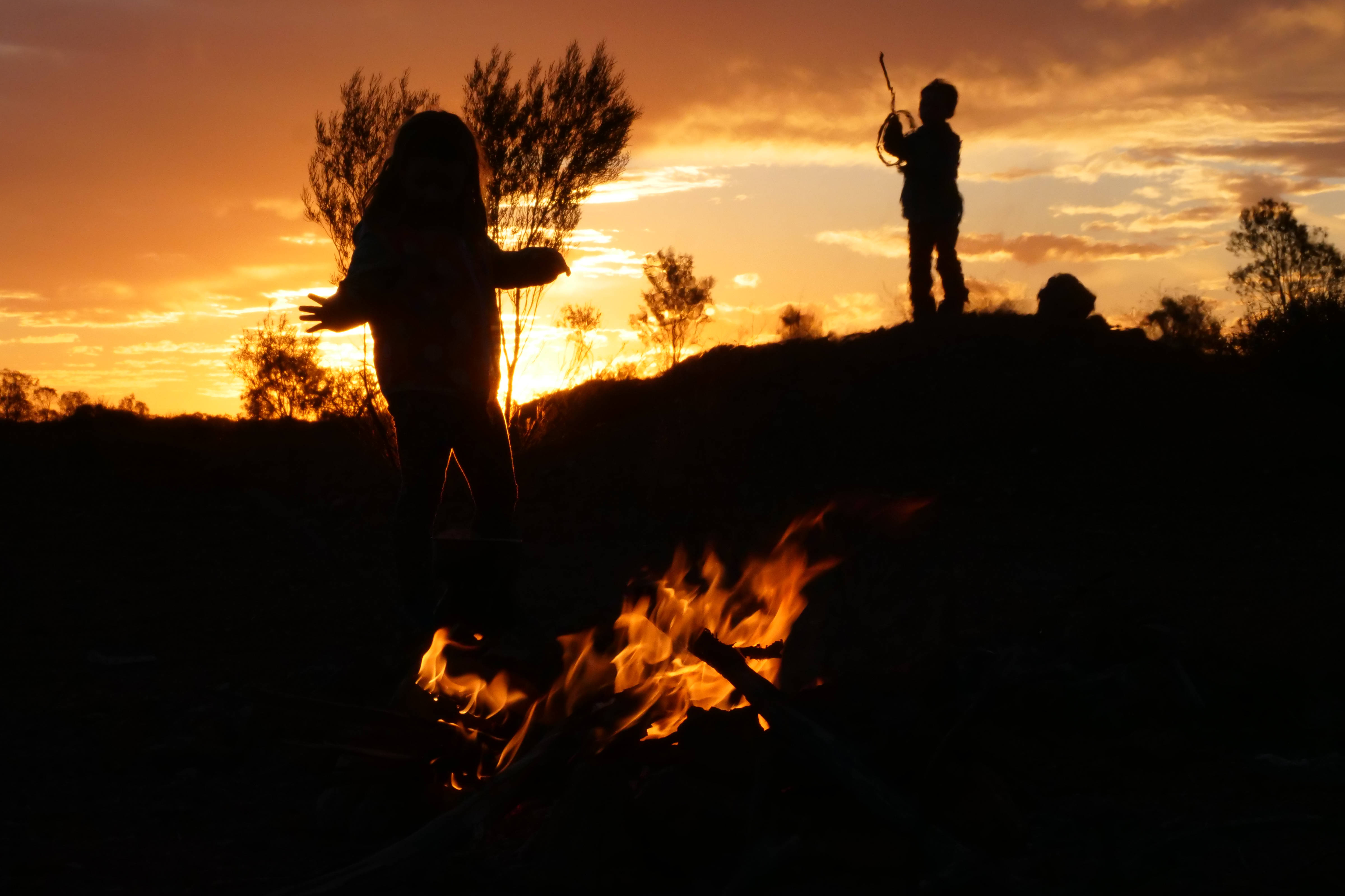 Silhouette of girl and boy with fire in foreground and sunset in background.